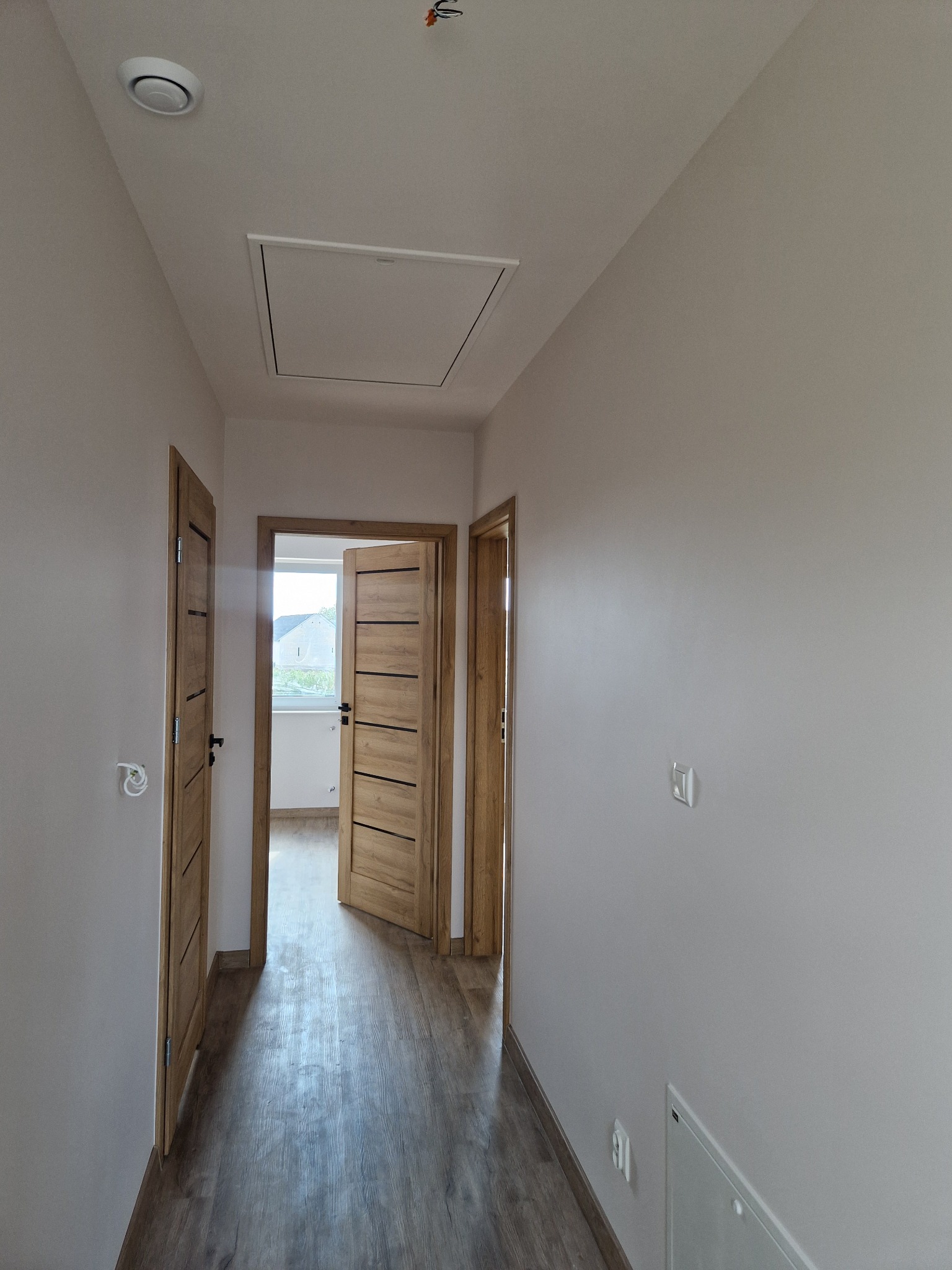 Korytarz w jasnych barwach z drewnopodobną podłogą, widoczne dwie drewniane drzwi z poziomymi intarsjami, jedna z nich lekko otwarta, sufit z klapą rewizyjną.