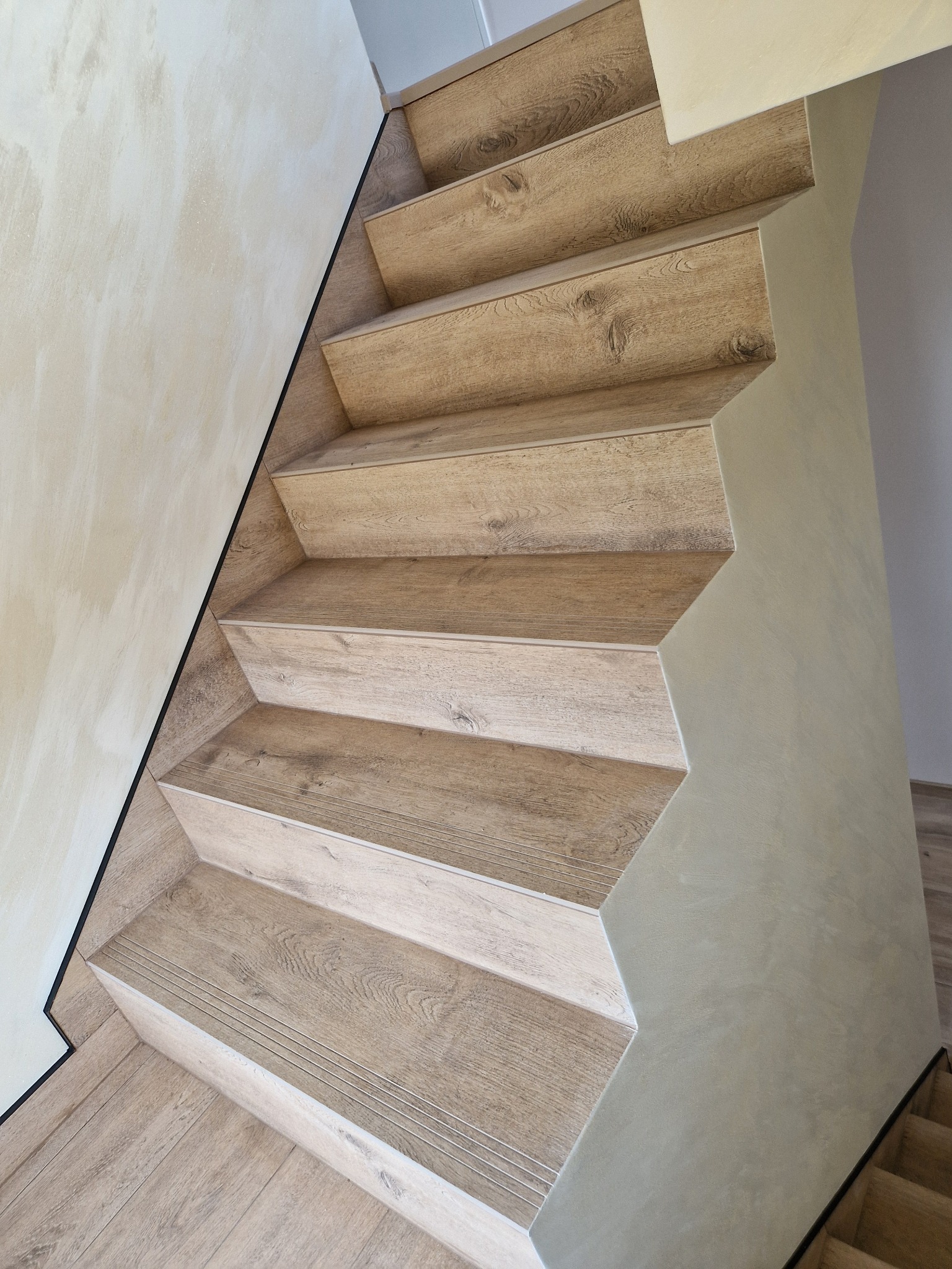 Schody wewnętrzne wykończone panelami imitującymi drewno, z czarną listwą przyścienną i ścianą pokrytą dekoracyjnym tynkiem.