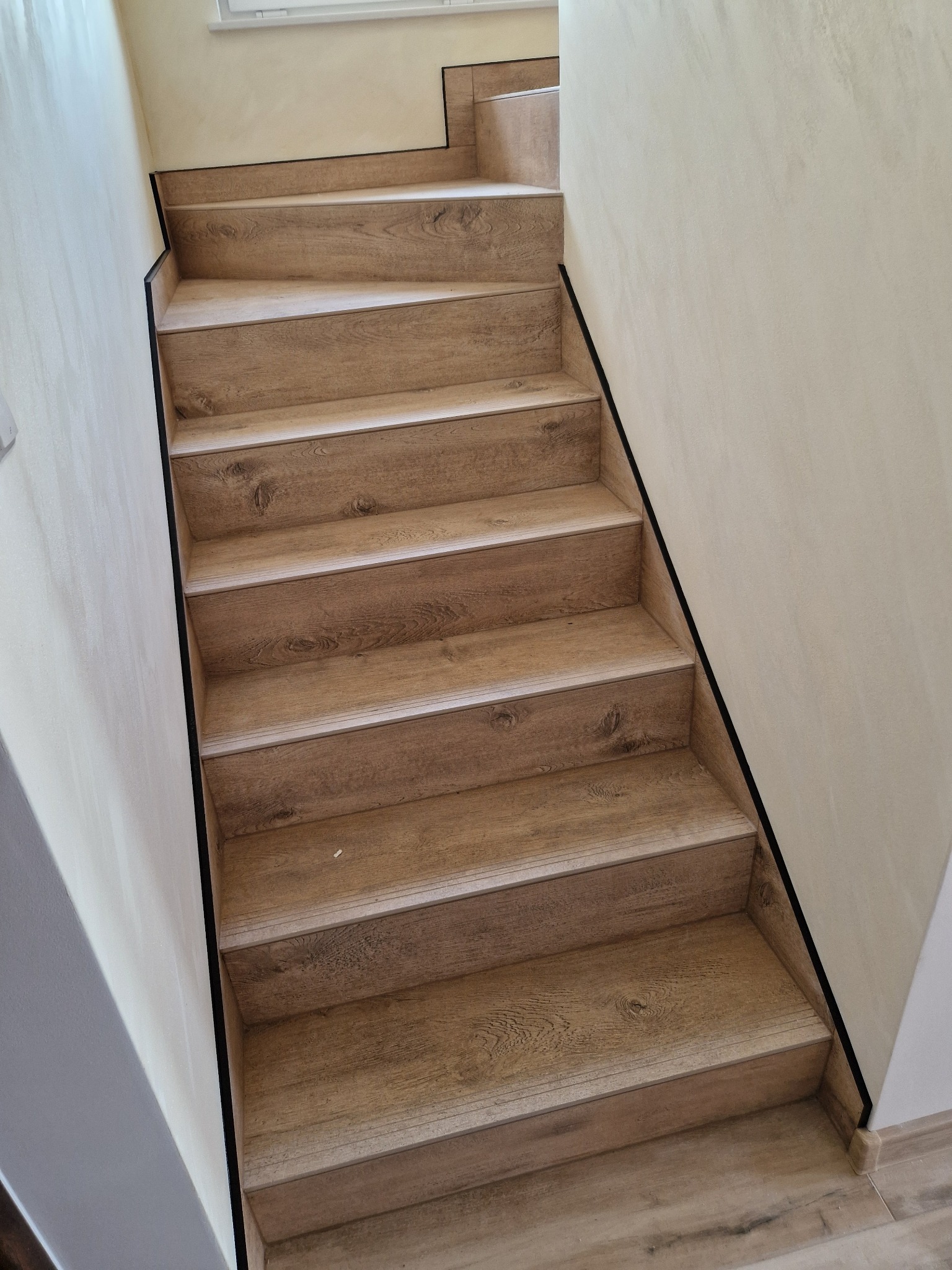 Schody wykończone panelami imitującymi drewno, z czarnymi listwami przypodłogowymi, ściany w jasnym odcieniu beżu z delikatną fakturą.