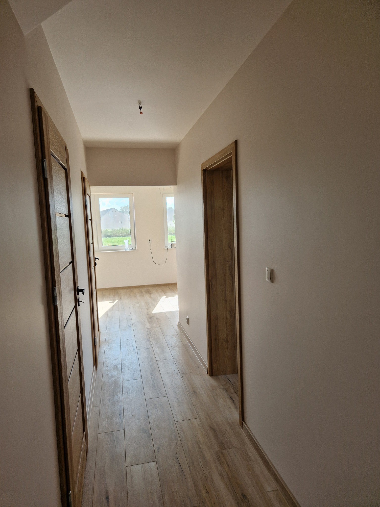 Wykończony korytarz z drewnopodobną podłogą, beżowymi ścianami i drewnianymi drzwiami z czarnymi klamkami, widok w głąb z dwoma oknami na końcu.
