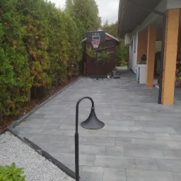 Ułożona szara kostka brukowa tworząca chodnik obok żywopłotu z tui, z lampą ogrodową w stylu retro na pierwszym planie i koszem do koszykówki w tle.