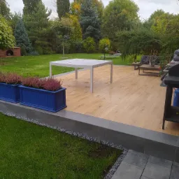 Nowoczesny taras z drewnianą podłogą, białym stołem i niebieskimi donicami z wrzosami, otoczony zielonym trawnikiem i drzewami.