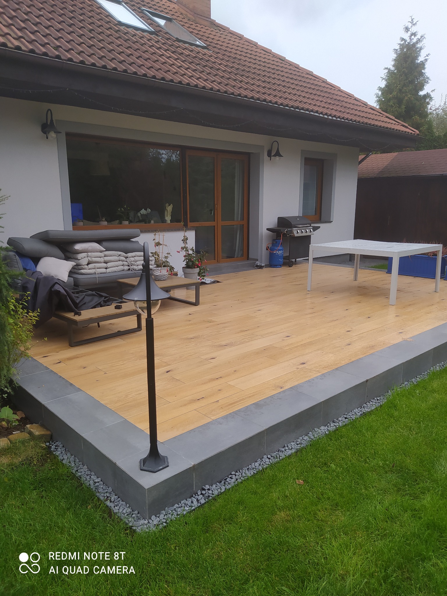 Drewniany taras z szarą obwódką z płytek, otoczony trawnikiem i kamieniami ozdobnymi, z meblami ogrodowymi, grillem i lampą stojącą, widok na dom z brązowym dachem.