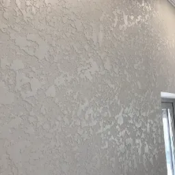 Teksturowana ściana w jasnym odcieniu szarości z dekoracyjnym efektem przypominającym nieregularne, delikatne wgłębienia i wypukłości, oświetlona z góry, z fragmentem okna po prawej stronie kadru.