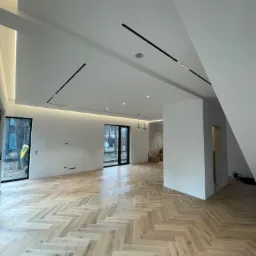 Jasny, przestronny pokój z parkietem ułożonym w jodełkę, białymi ścianami, czarnymi listwami oświetleniowymi na suficie i dużymi oknami z widokiem na zewnątrz. W tle widoczna surowa ściana...