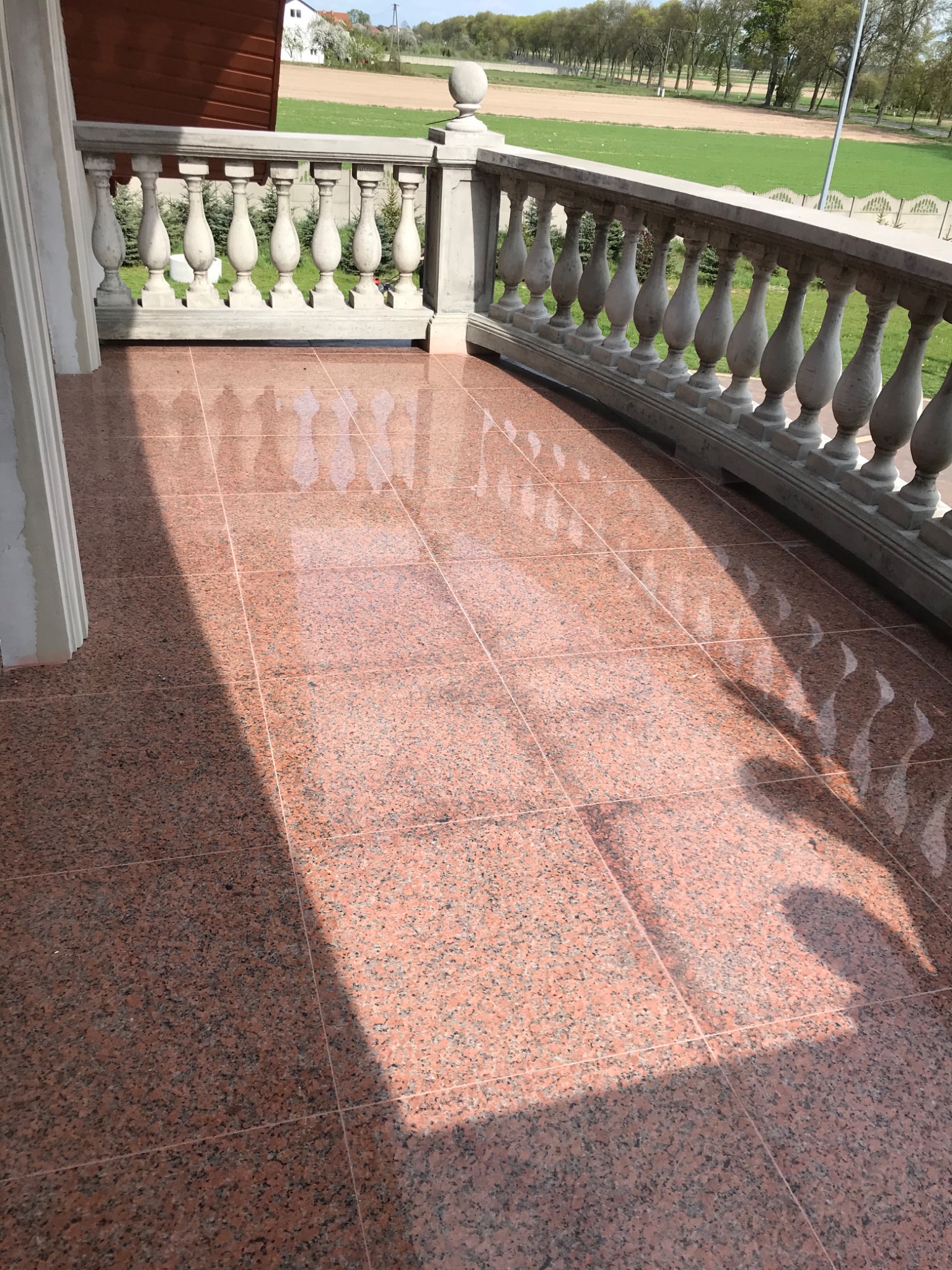 Balkon wyłożony błyszczącymi, różowo-szarymi płytkami granitowymi, z klasyczną białą balustradą i widokiem na zielone pole w słoneczny dzień.