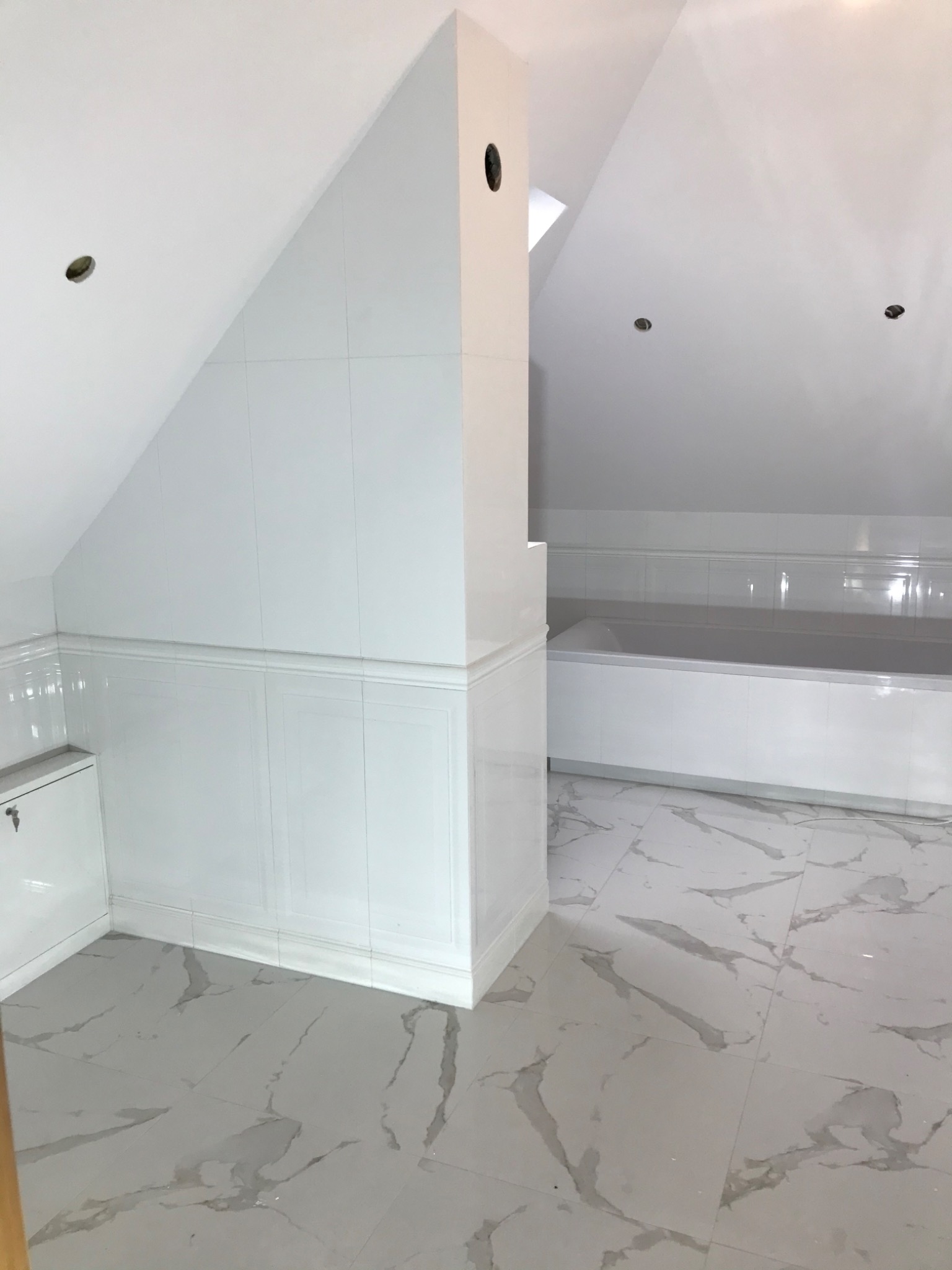 Wykończona łazienka na poddaszu z białą glazurą na ścianach i podłodze imitującą marmur, widoczna wanna i zabudowa.