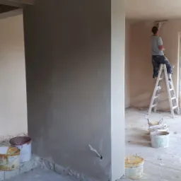 Nakładanie gładzi gipsowej na ścianę w nowym budynku, widoczny pracownik na drabinie, wiaderka z zaprawą i surowy stan wykończenia wnętrza.
