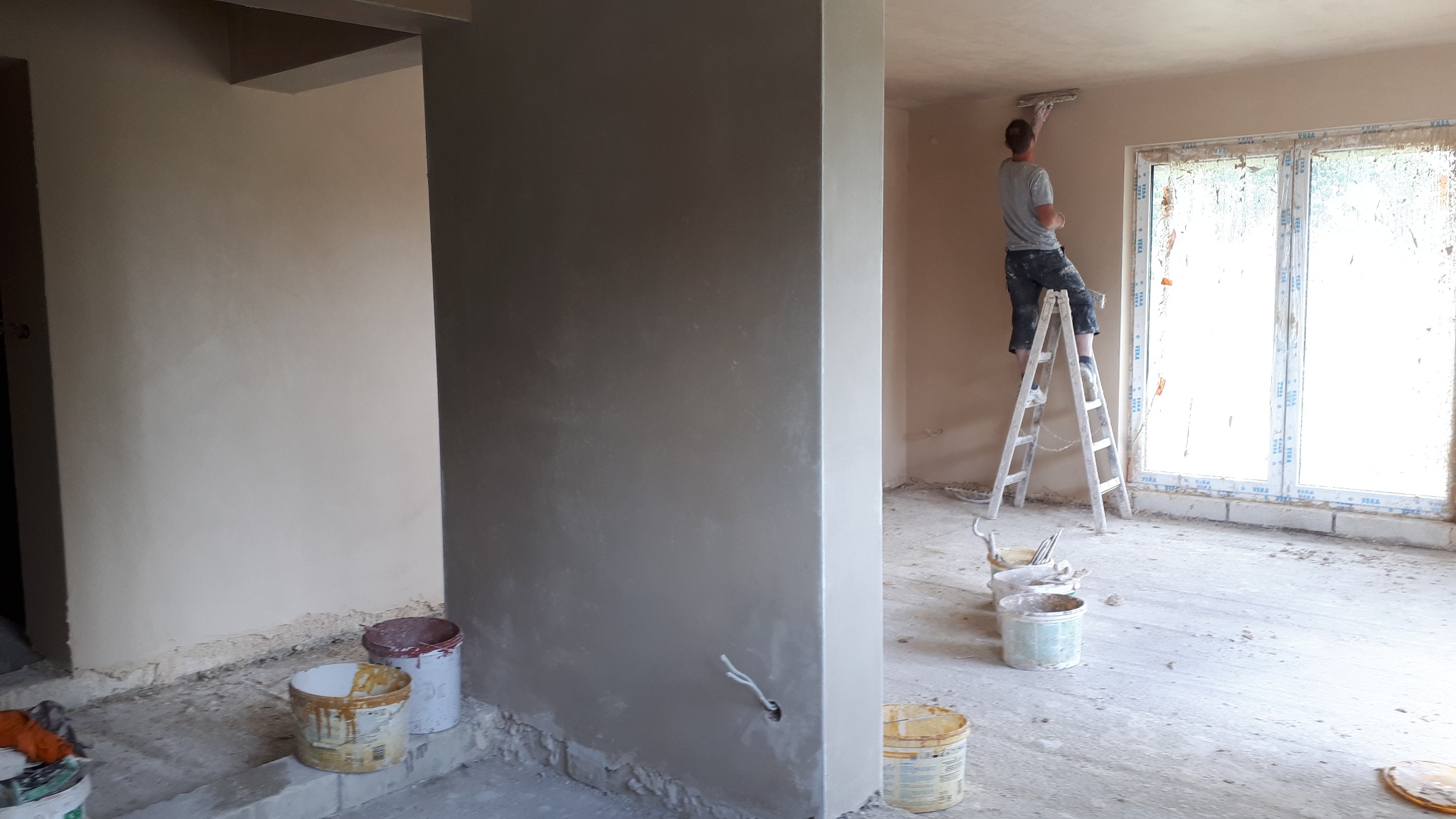 Nakładanie gładzi gipsowej na ścianę w nowym budynku, widoczny pracownik na drabinie, wiaderka z zaprawą i surowy stan wykończenia wnętrza.