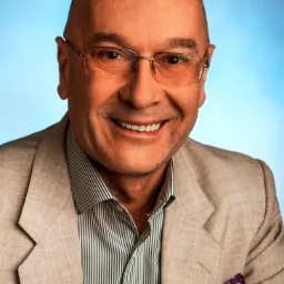 Portret uśmiechniętego mężczyzny w okularach, beżowej marynarce i koszuli w paski na tle jasnoniebieskim.