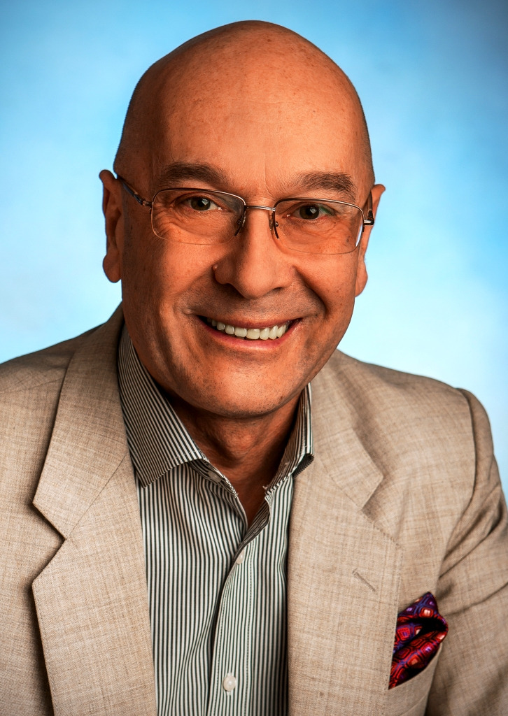 Portret uśmiechniętego mężczyzny w okularach, beżowej marynarce i koszuli w paski na tle jasnoniebieskim.