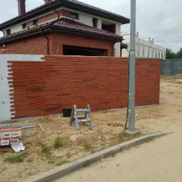 Murowanie ogrodzenia z cegły klinkierowej, widoczny fragment ściany z surowego betonu, drabina aluminiowa i paczki cegieł Kingsize obok, w tle dom jednorodzinny z czerwoną dach&oacute;wką.