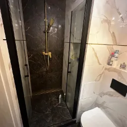 Wykończona łazienka z kabiną prysznicową z czarnymi profilami i złotą armaturą na tle marmurowych płytek.