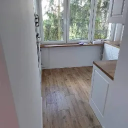 Wnętrze kuchni po remoncie: świeżo pomalowane ściany w odcieniu bieli, drewnopodobne płytki na podłodze i widok na zieleń za oknem.