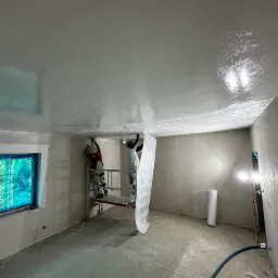 Ekipa na rusztowaniu wygładza sufit w pomieszczeniu, w tle okno z folią ochronną i lampa doświetlająca, podłoga surowa, widoczne narzędzia i materiały.