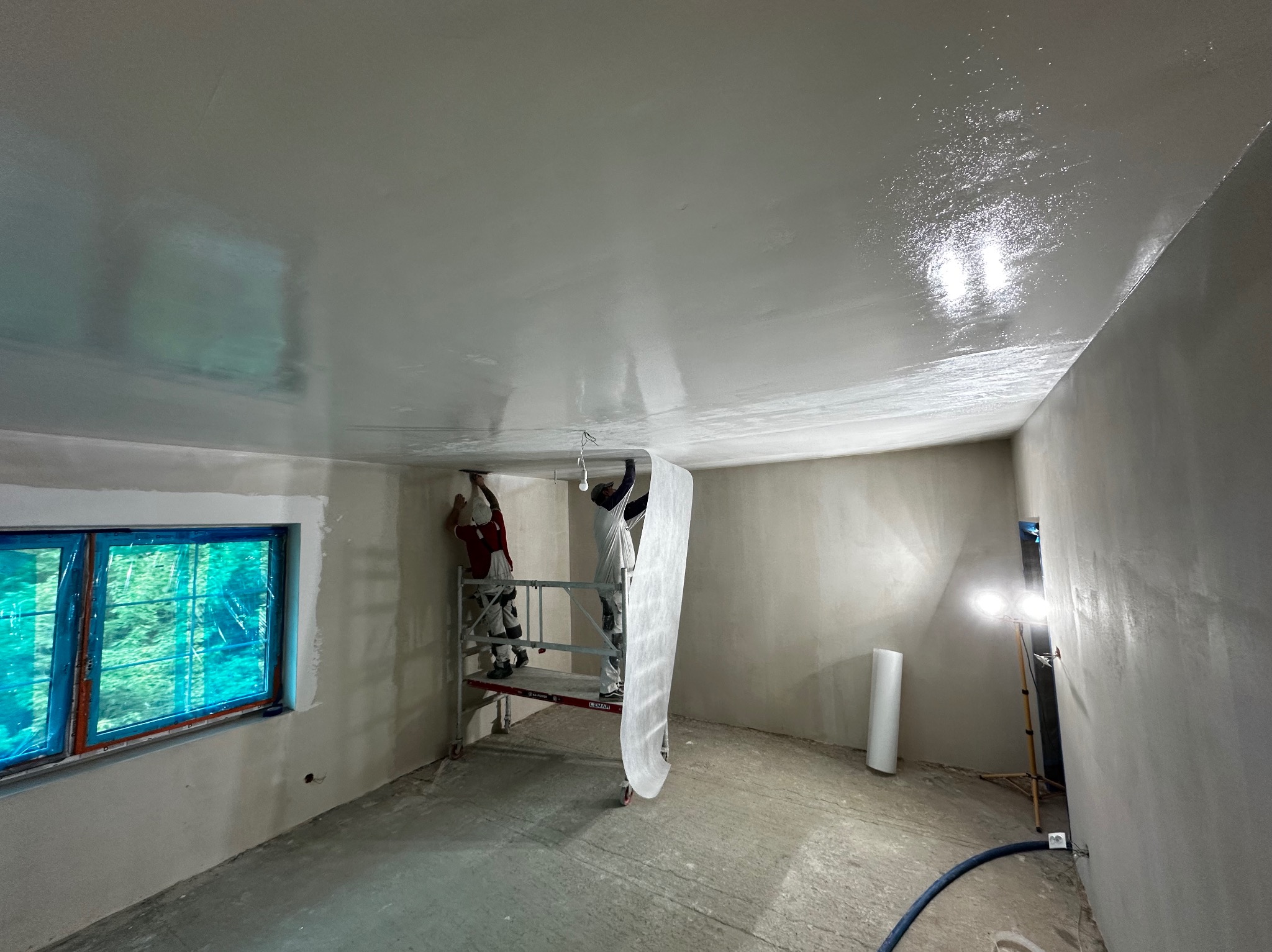 Ekipa na rusztowaniu wygładza sufit w pomieszczeniu, w tle okno z folią ochronną i lampa doświetlająca, podłoga surowa, widoczne narzędzia i materiały.