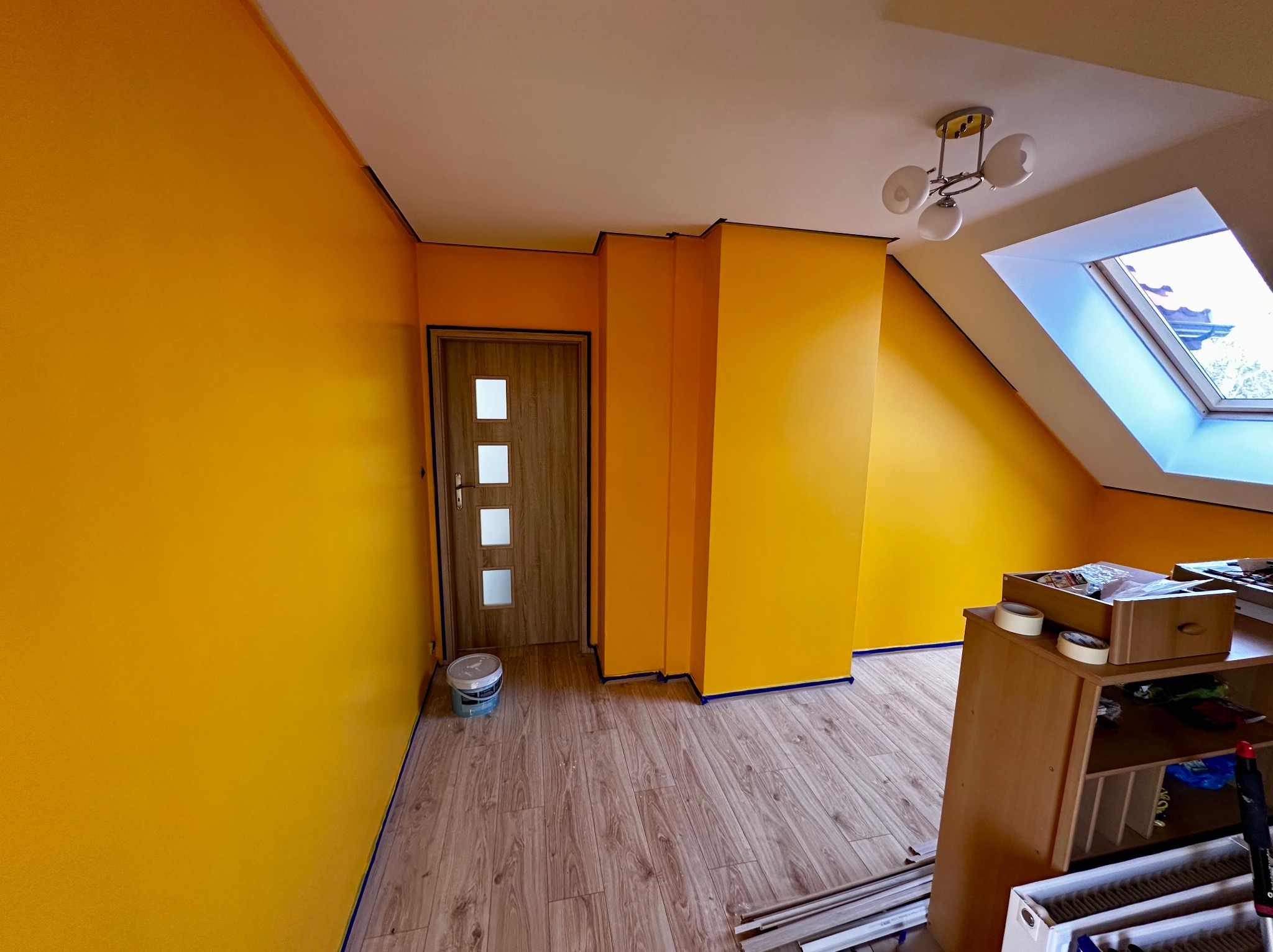 Żółte ściany świeżo po malowaniu w pokoju na poddaszu. Widoczna drewniana podłoga, białe okno dachowe i drzwi. Użyto taśmy malarskiej dla precyzji.