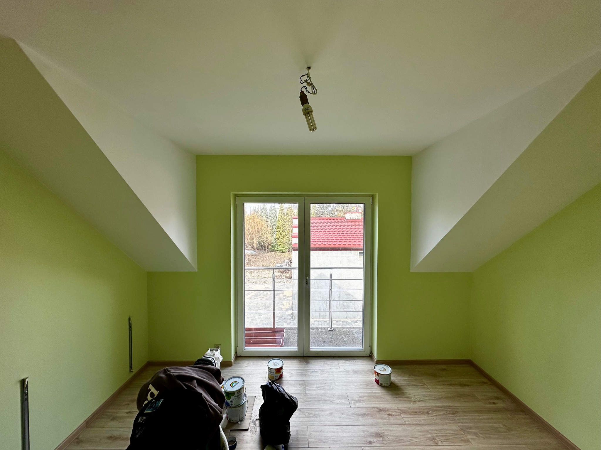 Pomalowane na zielono poddasze z oknem balkonowym. Widoczne puszki z farbą na drewnianej podłodze. Biały sufit i skosy.