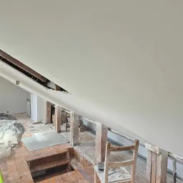 Skośny sufit po nałożeniu gładzi, widoczne drewniane elementy konstrukcyjne, fragment podłogi z otworem i prowizoryczne schodki, krzesło robocze w tle.