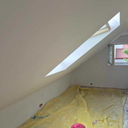 LA CASA BALTICA DAMIAN SULAK - Poddasze po świeżym pomalowaniu na biało, zabezpieczone folią malarską, widoczne okno dachowe i zwykłe, na podłodze leżą buty i ubrania.