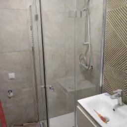 Wykończona kabina prysznicowa z elementami chromowanymi, umywalką i baterią. Na umywalce leży śrubokręt. Tło stanowią płytki w beżach i geometryczne wzory.