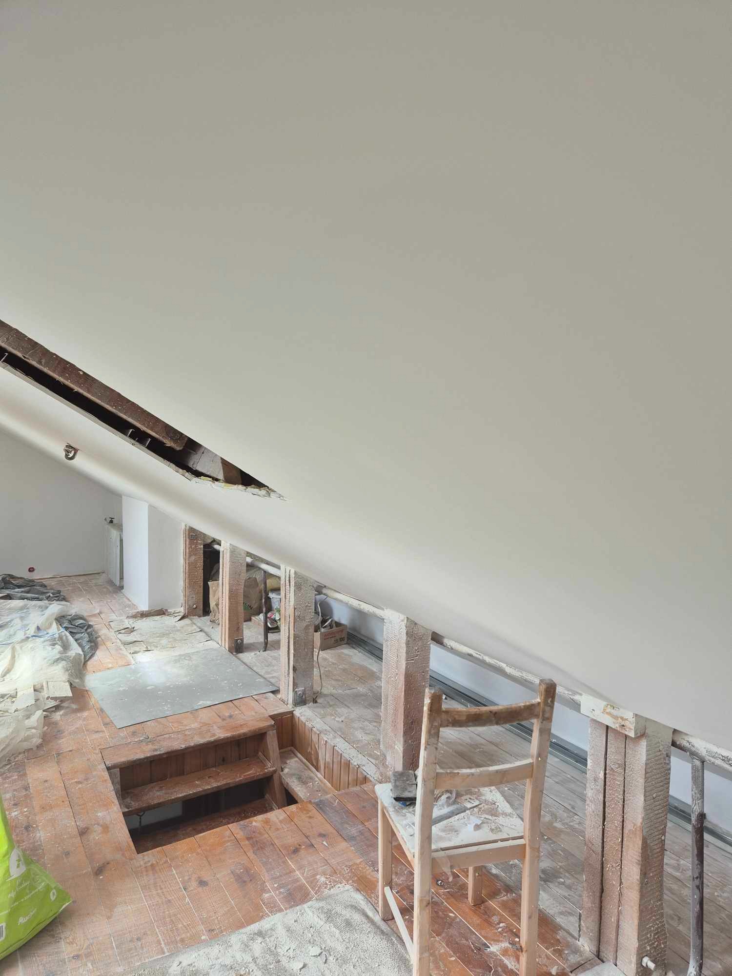 Skośny sufit po nałożeniu gładzi, widoczne drewniane elementy konstrukcyjne, fragment podłogi z otworem i prowizoryczne schodki, krzesło robocze w tle.