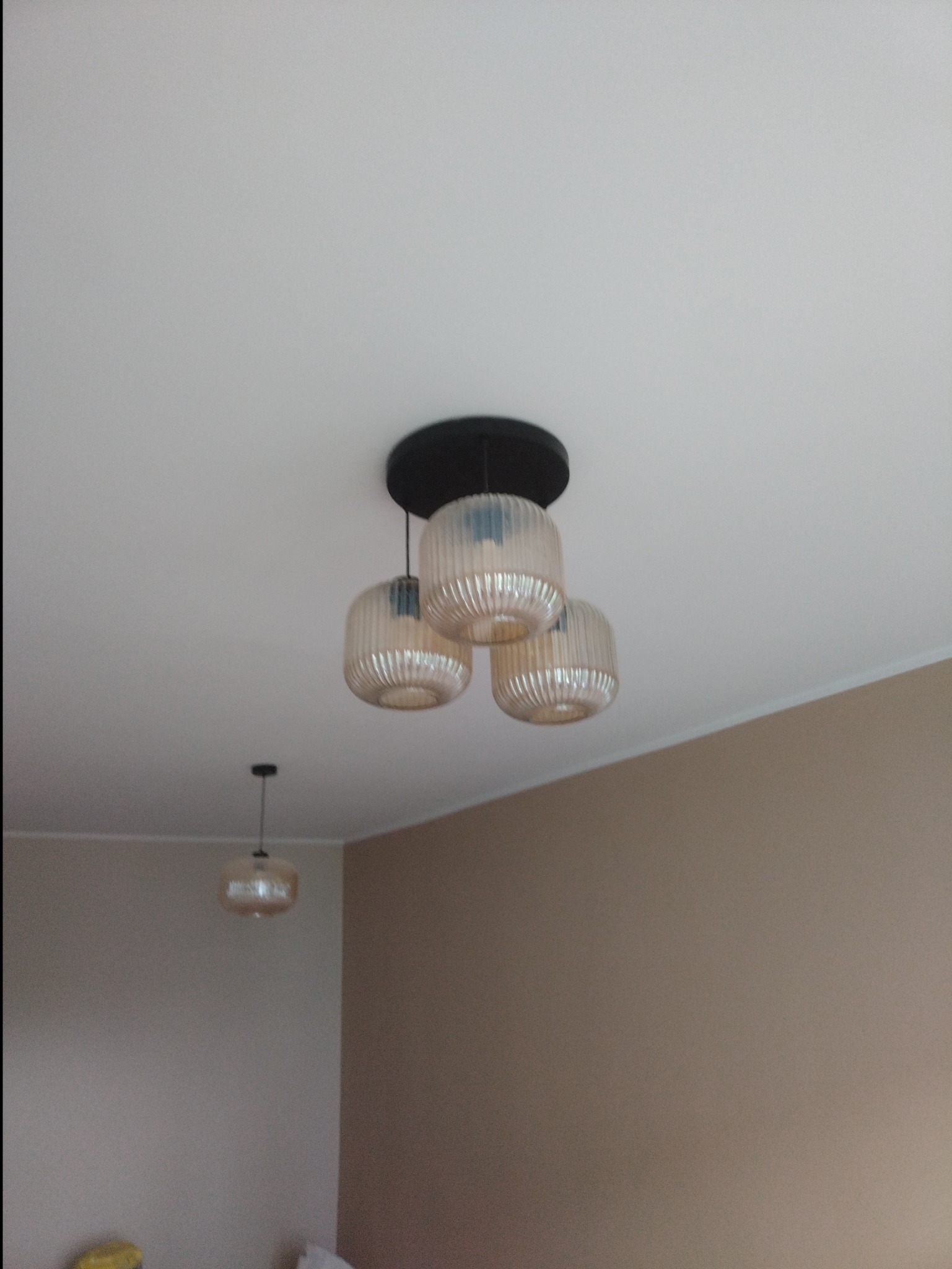 Beżowa lampa sufitowa z trzema kloszami w prążki, na tle białego sufitu i beżowych ścian. Widoczna druga, mniejsza lampa w tle.