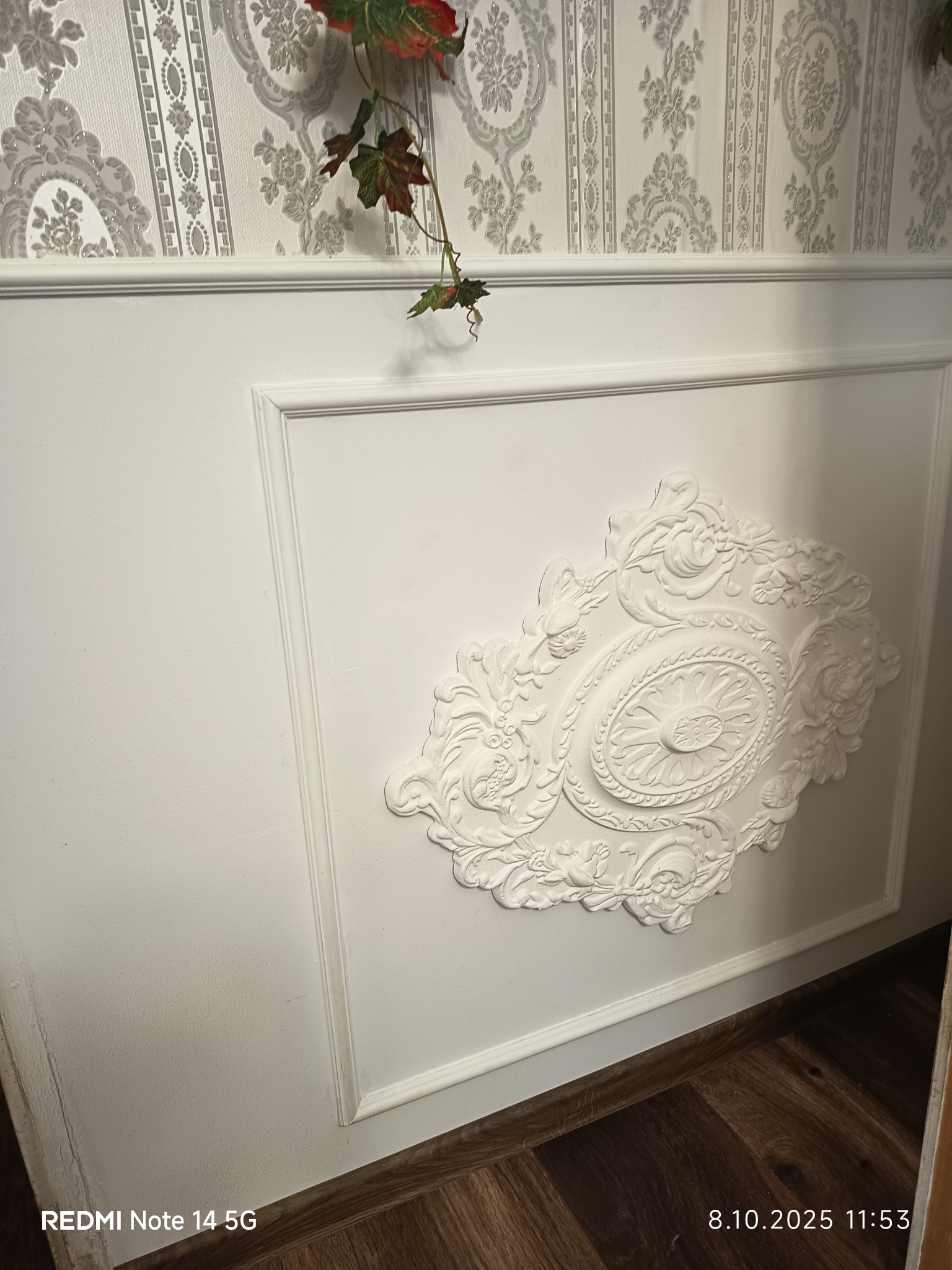 Biała ściana z dekoracyjnym panelem i sztukaterią, nad nią tapeta w ornamenty. Element wykończenia wnętrza.