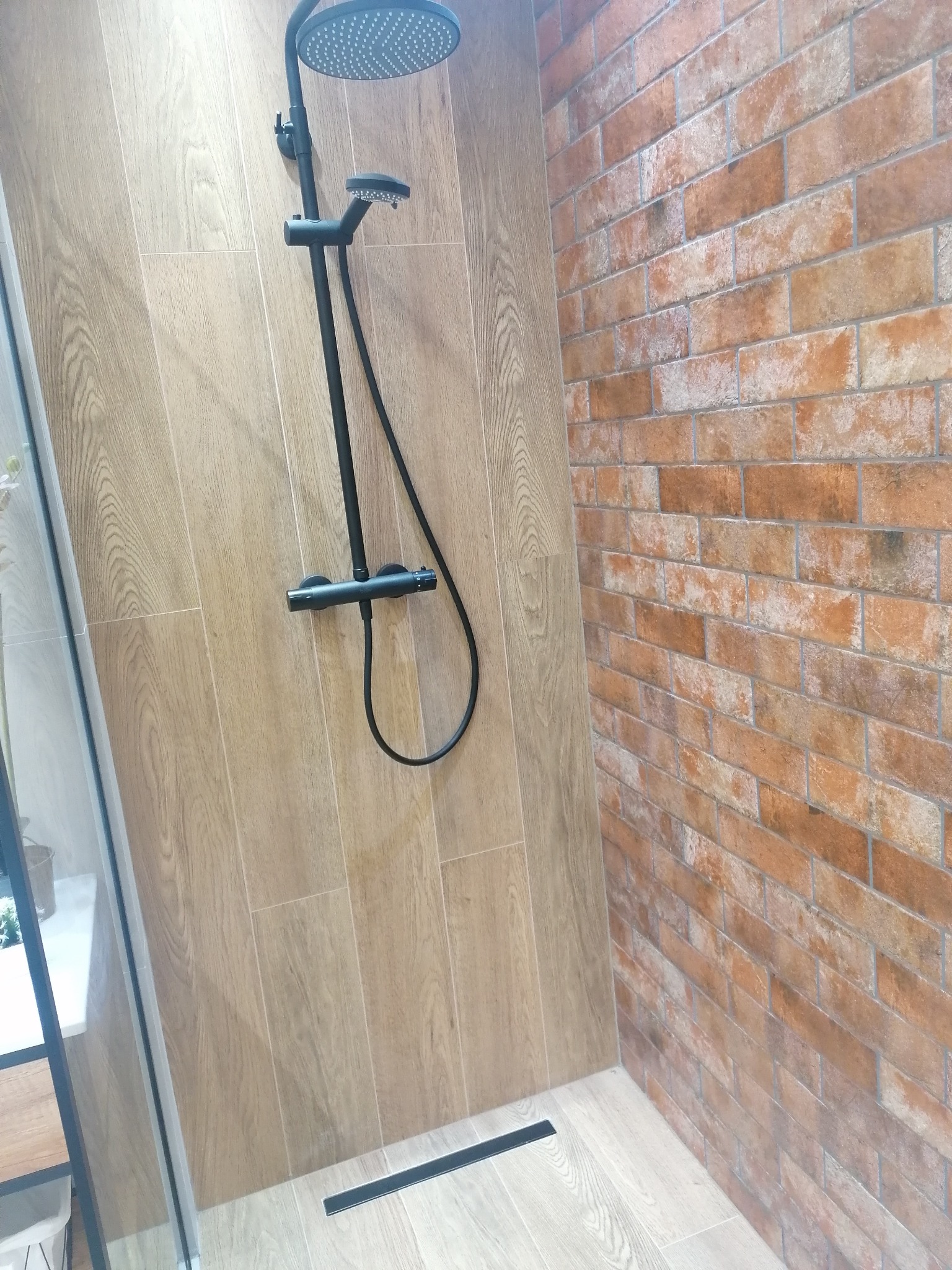 Nowoczesna kabina prysznicowa z deszczownicą, wykończona płytkami imitującymi drewno i cegłę, z czarnym odpływem liniowym i szklaną ścianką.