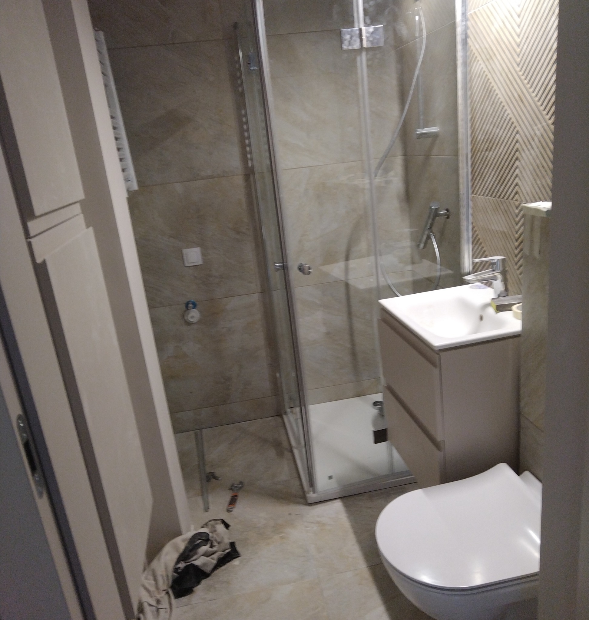 Jasna łazienka po remoncie: kabina prysznicowa ze szklanymi drzwiami, umywalka z szafką, toaleta. Na podłodze widoczne narzędzia i szmatka.