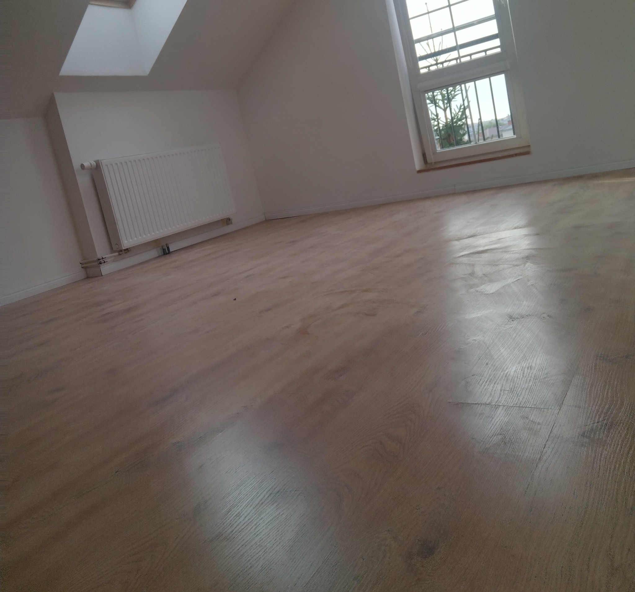 Jasne panele podłogowe w pokoju na poddaszu z oknem i grzejnikiem. Widoczne skosy i okno dachowe. Ukończony efekt układania paneli.