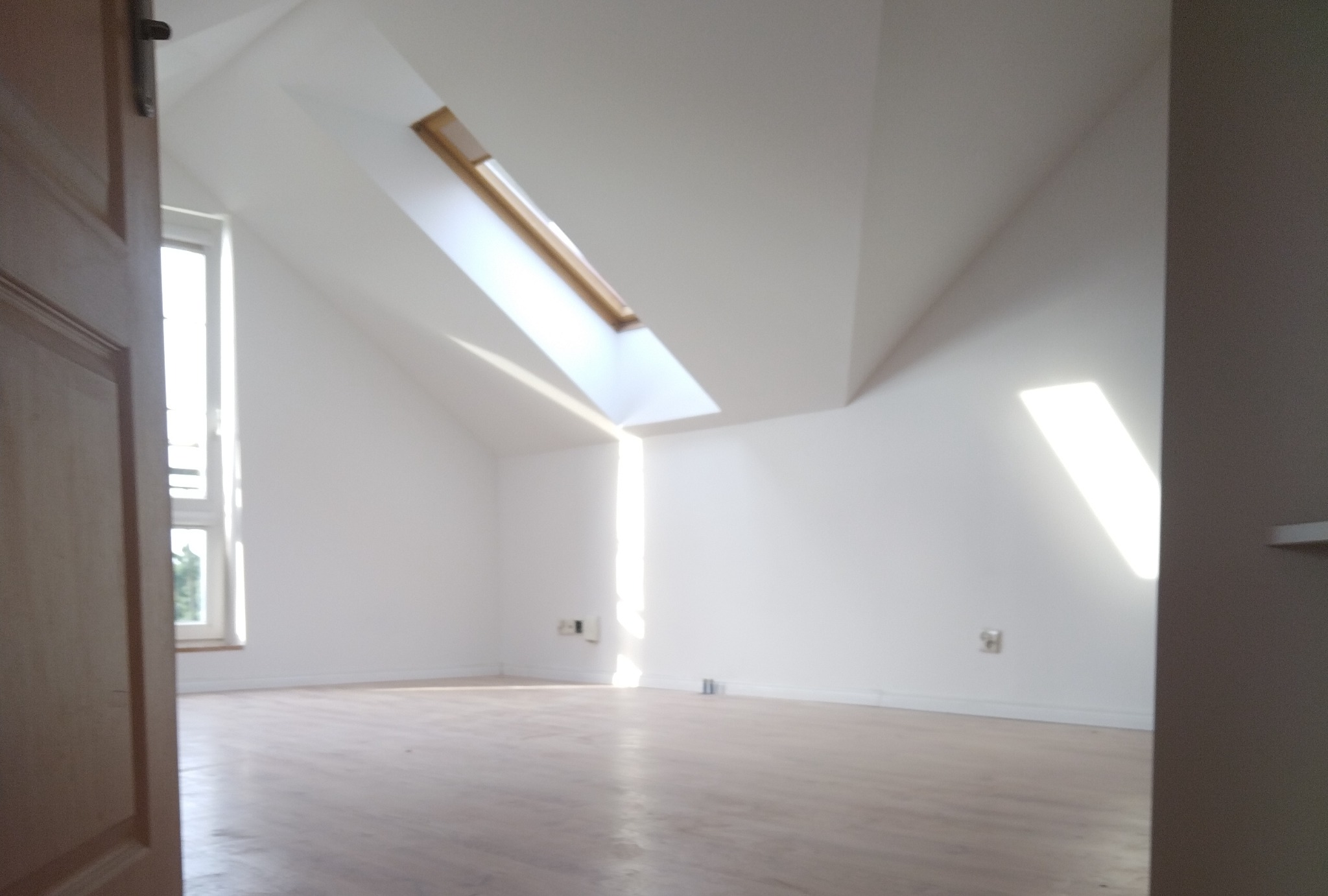 Jasne poddasze z oknem dachowym i drewnianą podłogą, świeżo po remoncie. Widok zza uchylonych drzwi na puste, pomalowane na biało wnętrze.