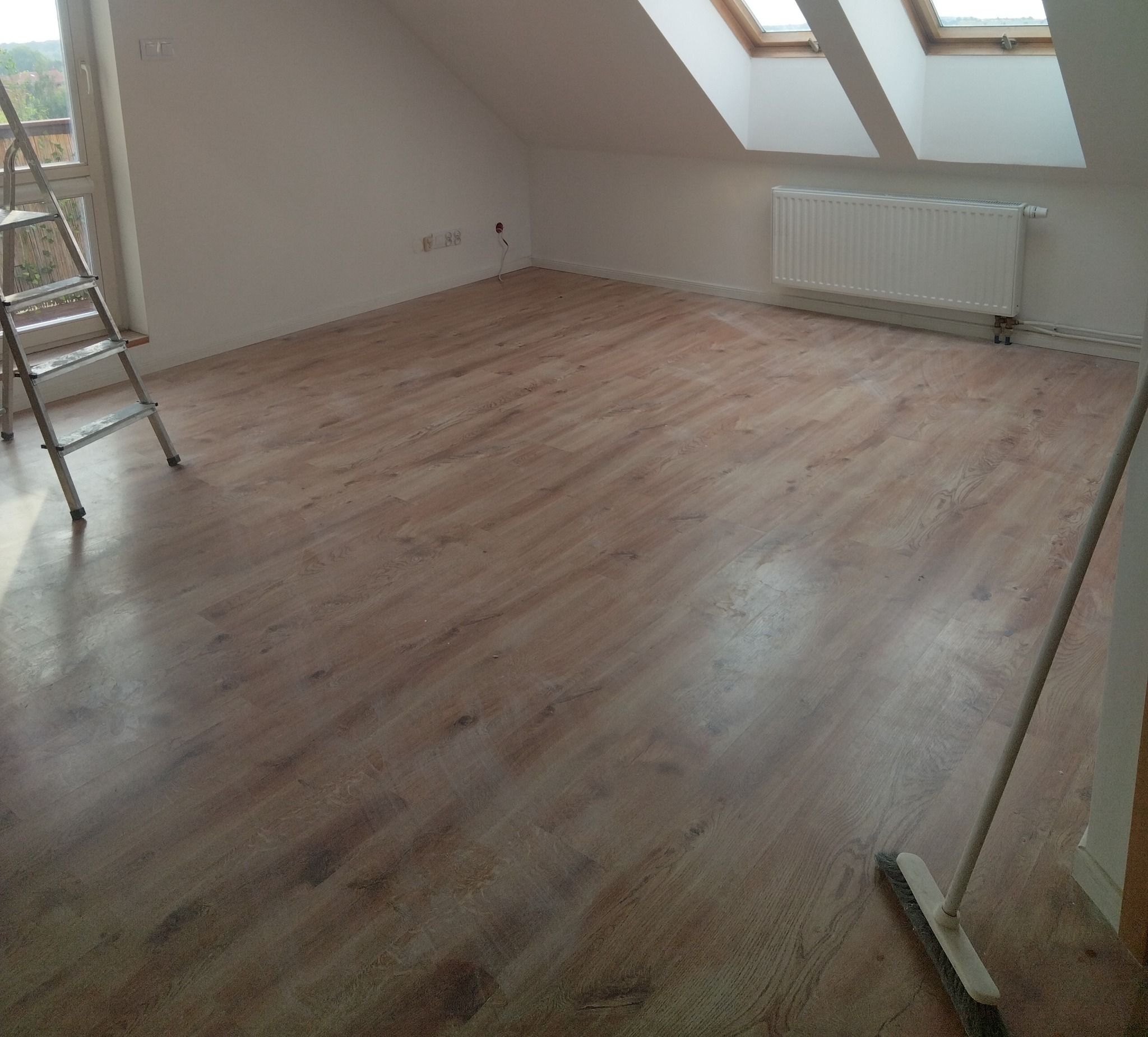 Jasne panele podłogowe w pokoju na poddaszu z oknami dachowymi. Widoczna drabina malarska i szczotka, sugerujące prace wykończeniowe.