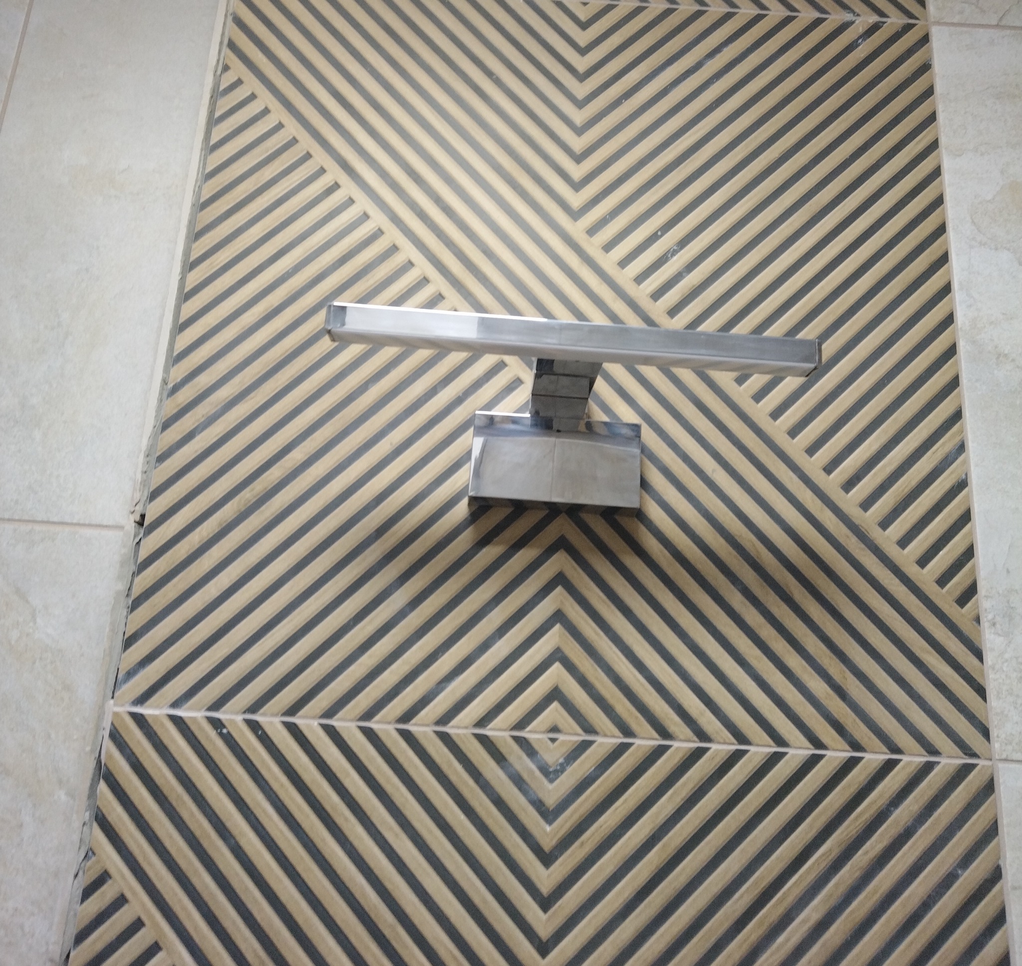 Ściana z dekoracyjną mozaiką w geometryczny wzór, oświetlona nowoczesną, srebrną lampą ścienną. Widoczne płytki na ścianie.