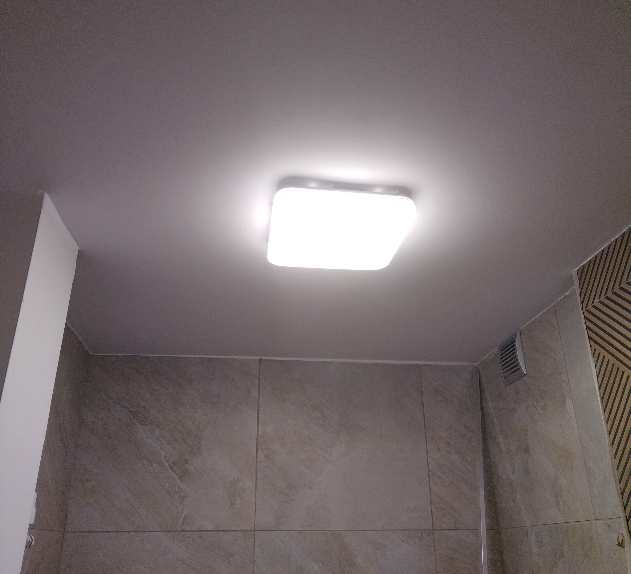 Wykończona łazienka z beżowymi płytkami i nowoczesną, kwadratową lampą sufitową LED. Widoczny fragment ściany z dekoracyjnym wzorem.