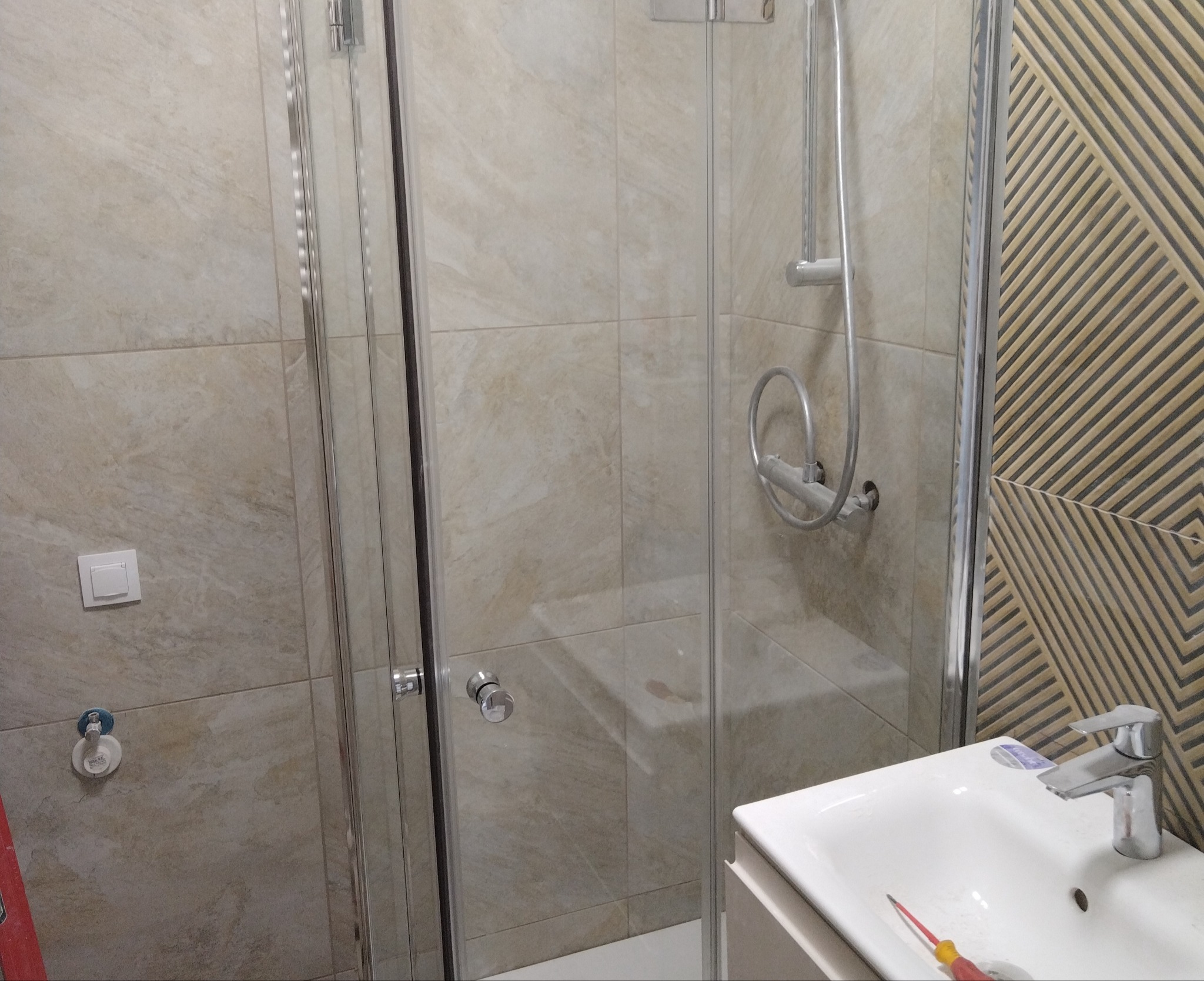 Wykończona łazienka z kabiną prysznicową, umywalką i ozdobnymi płytkami. Widoczne detale, takie jak bateria i przełącznik światła. Całość utrzymana w jasnej kolorystyce.