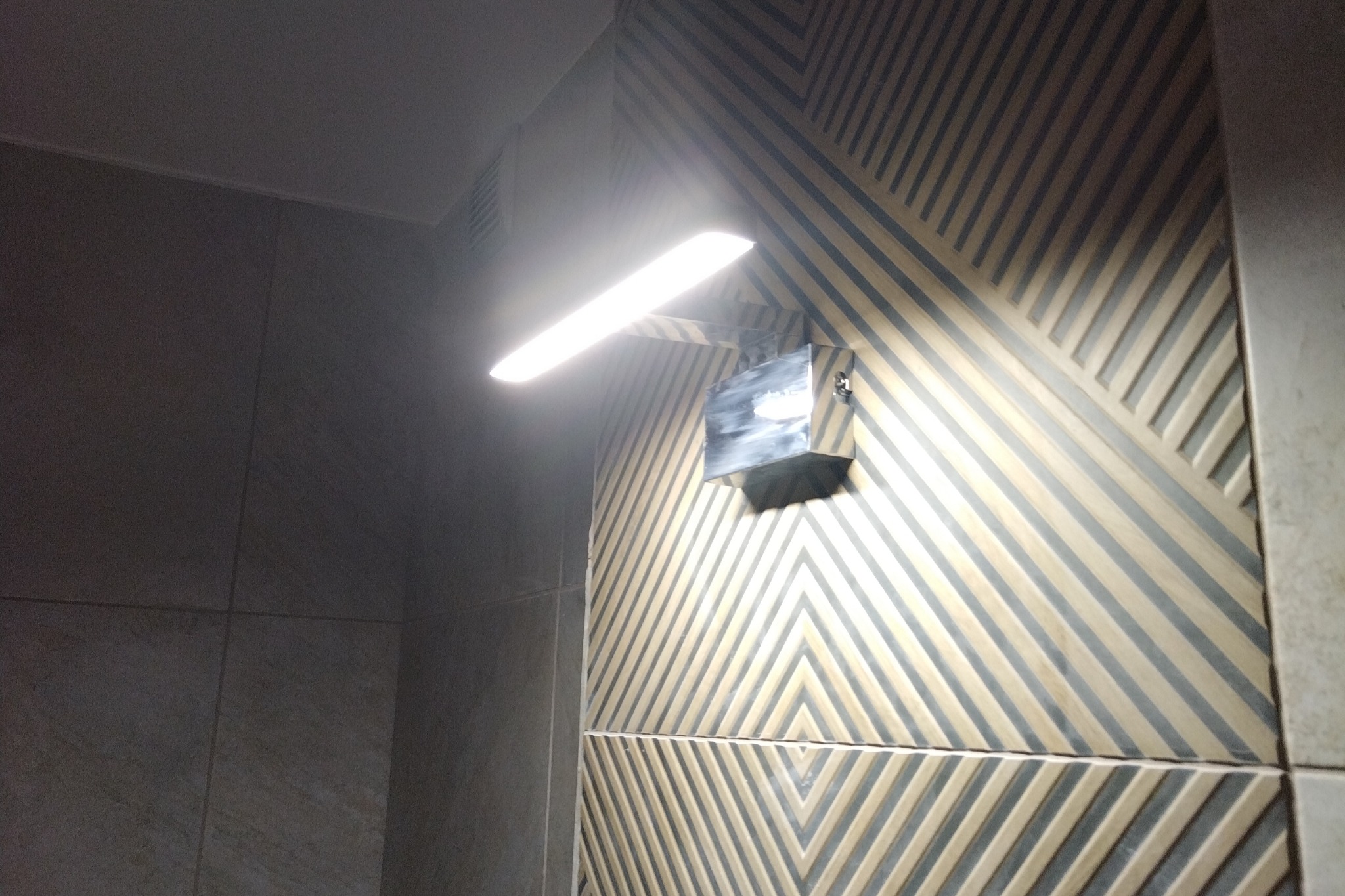 Świecąca lampa ścienna nad wzorzystą, beżowo-szarą ścianą z płytek w łazience. Nietypowy układ płytek tworzy geometryczny deseń.