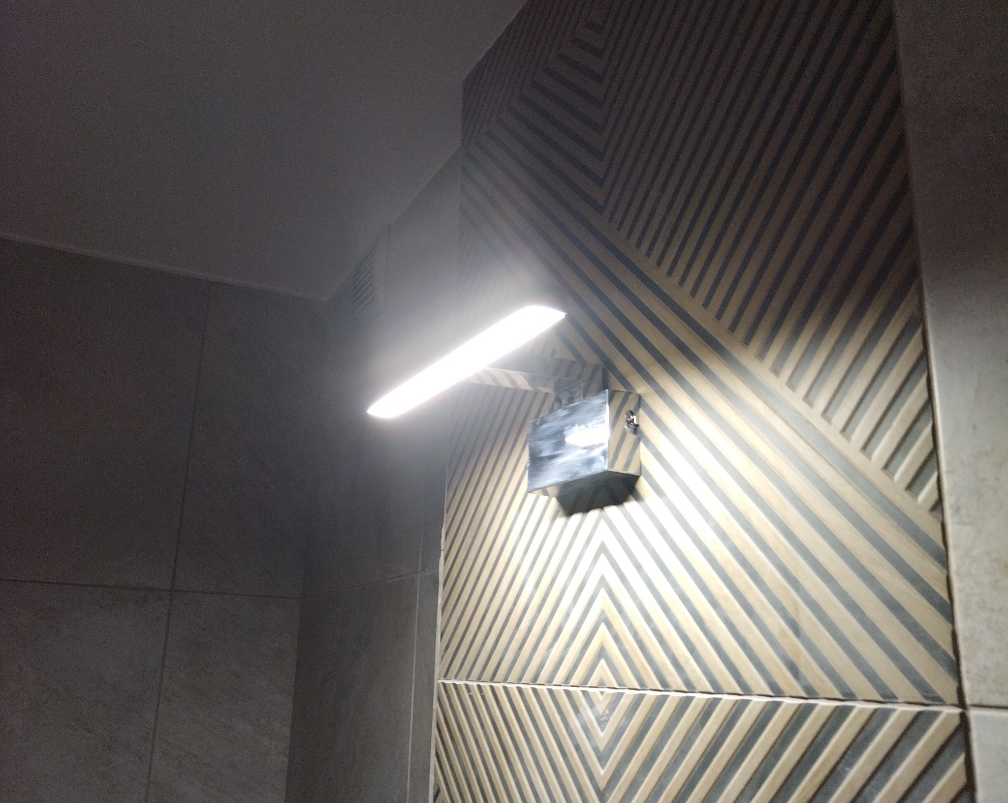 Świecąca lampa ścienna nad wzorzystą, wykończoną ścianą w łazience. Geometryczny deseń na płytkach, oświetlony jasnym światłem lampy.