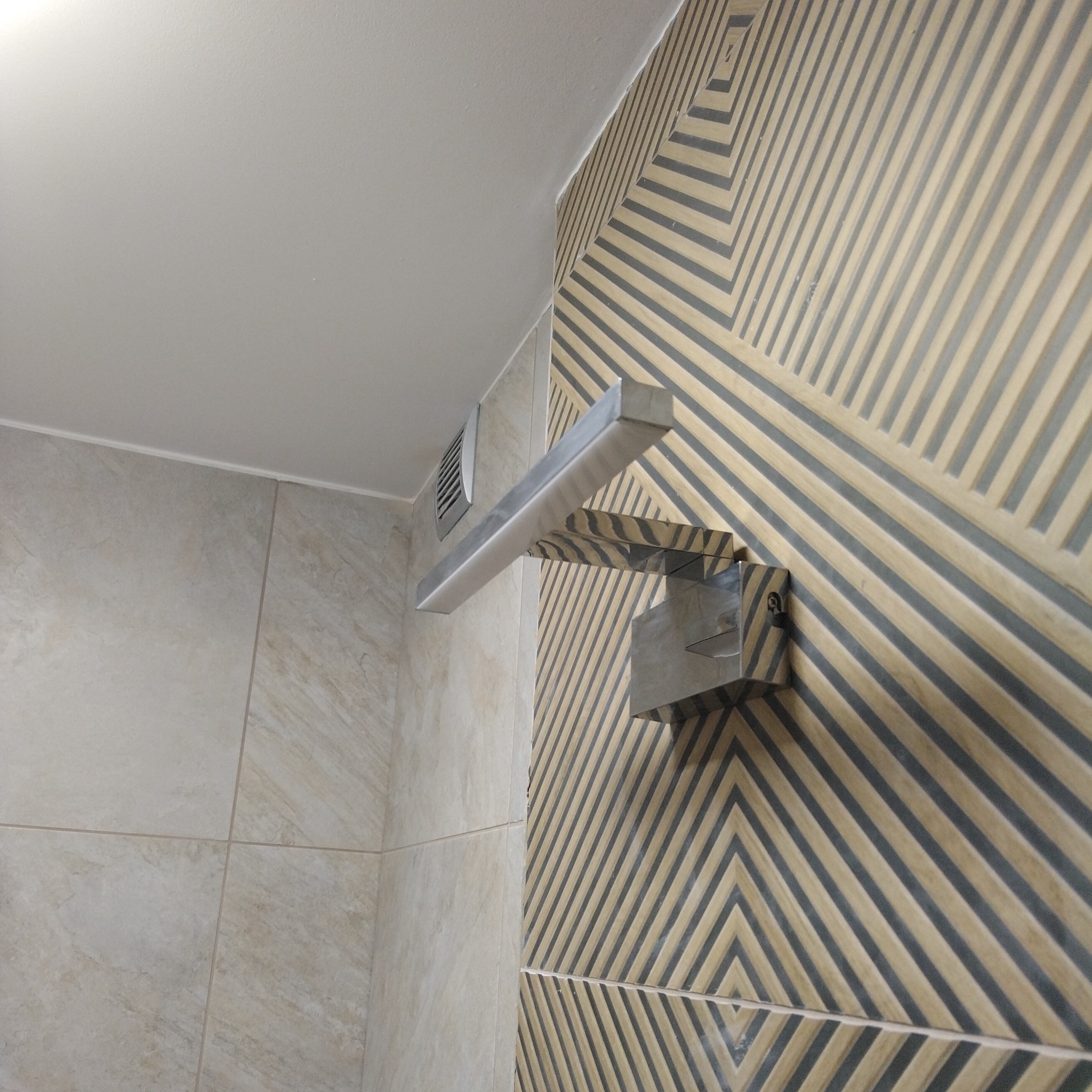 Wykończona łazienka z nowoczesną lampą nad lustrem, na ścianie płytki z geometrycznym wzorem. Widoczny fragment wentylacji i gładkie, jasne płytki na ścianie obok.