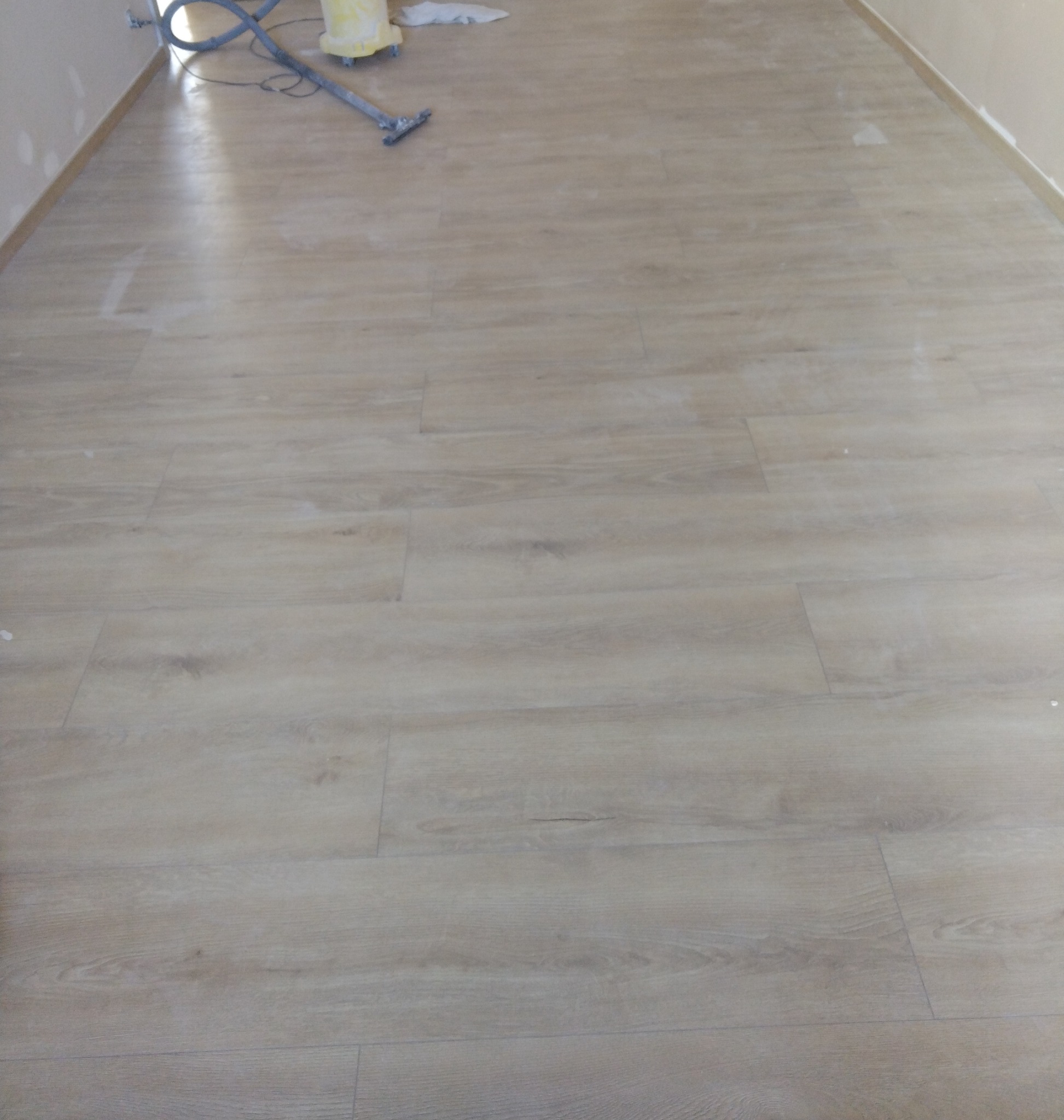 Jasne panele podłogowe ułożone w nowym wnętrzu, z widocznymi narzędziami do sprzątania w rogu. Minimalistyczny styl, świeżo po montażu.