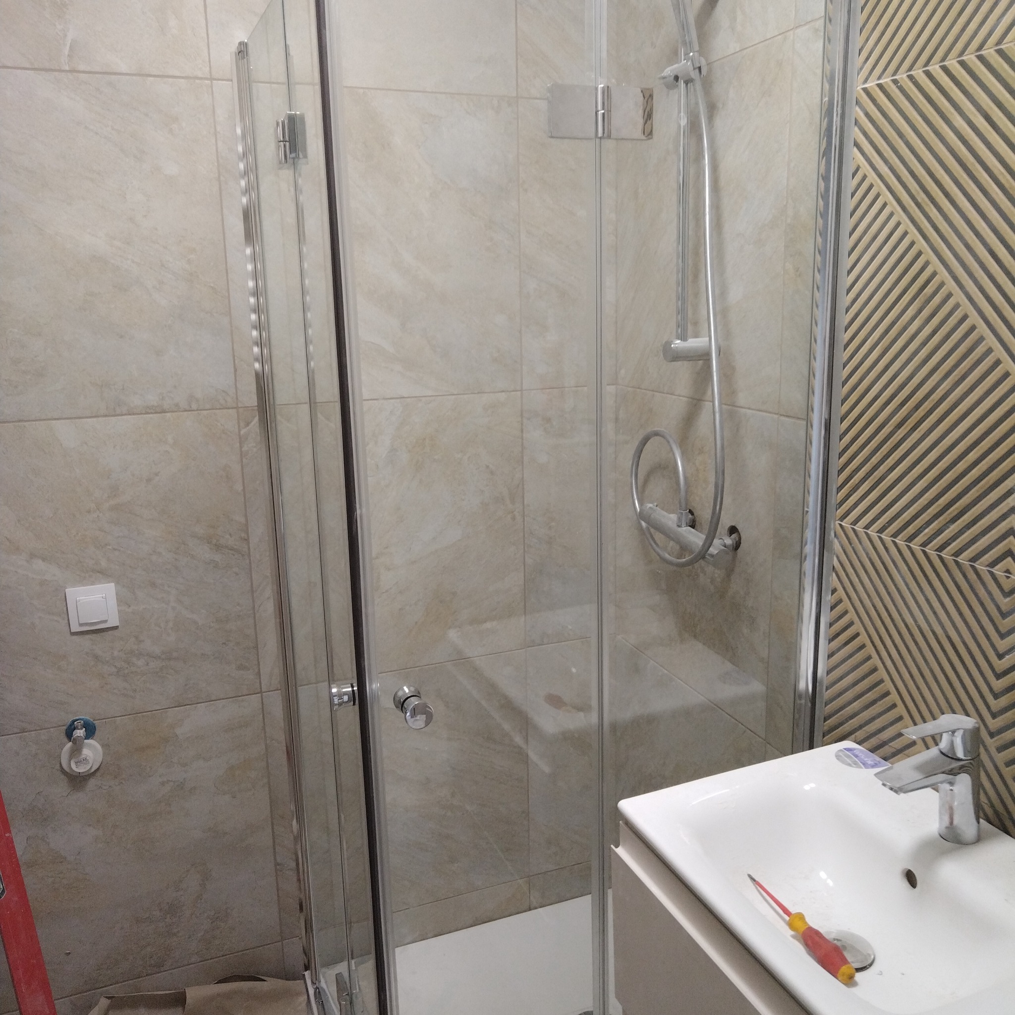 Wykończona kabina prysznicowa z elementami chromowanymi, umywalką i baterią. Na umywalce leży śrubokręt. Tło stanowią płytki w beżach i geometryczne wzory.