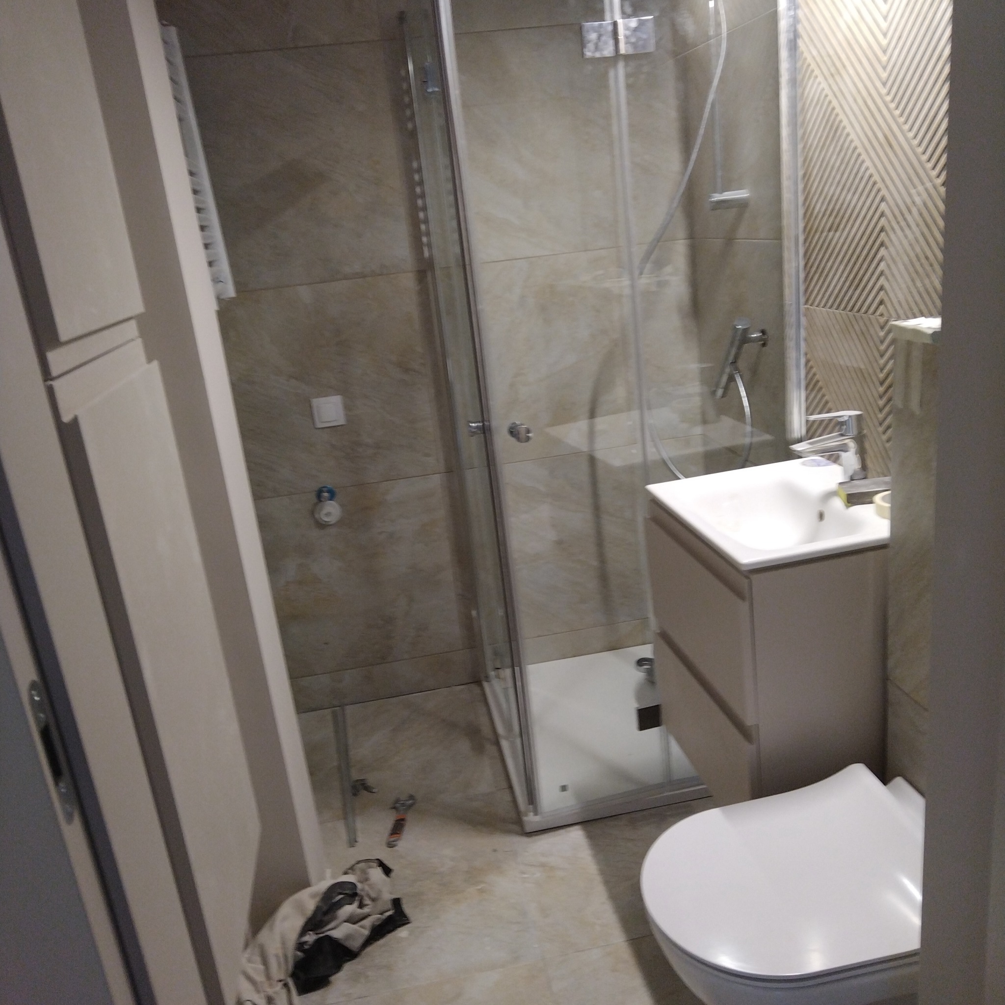 Wykończona łazienka z kabiną prysznicową, umywalką z szafką i toaletą. Na podłodze narzędzia i szmatka. Ściany wyłożone płytkami w odcieniach beżu.