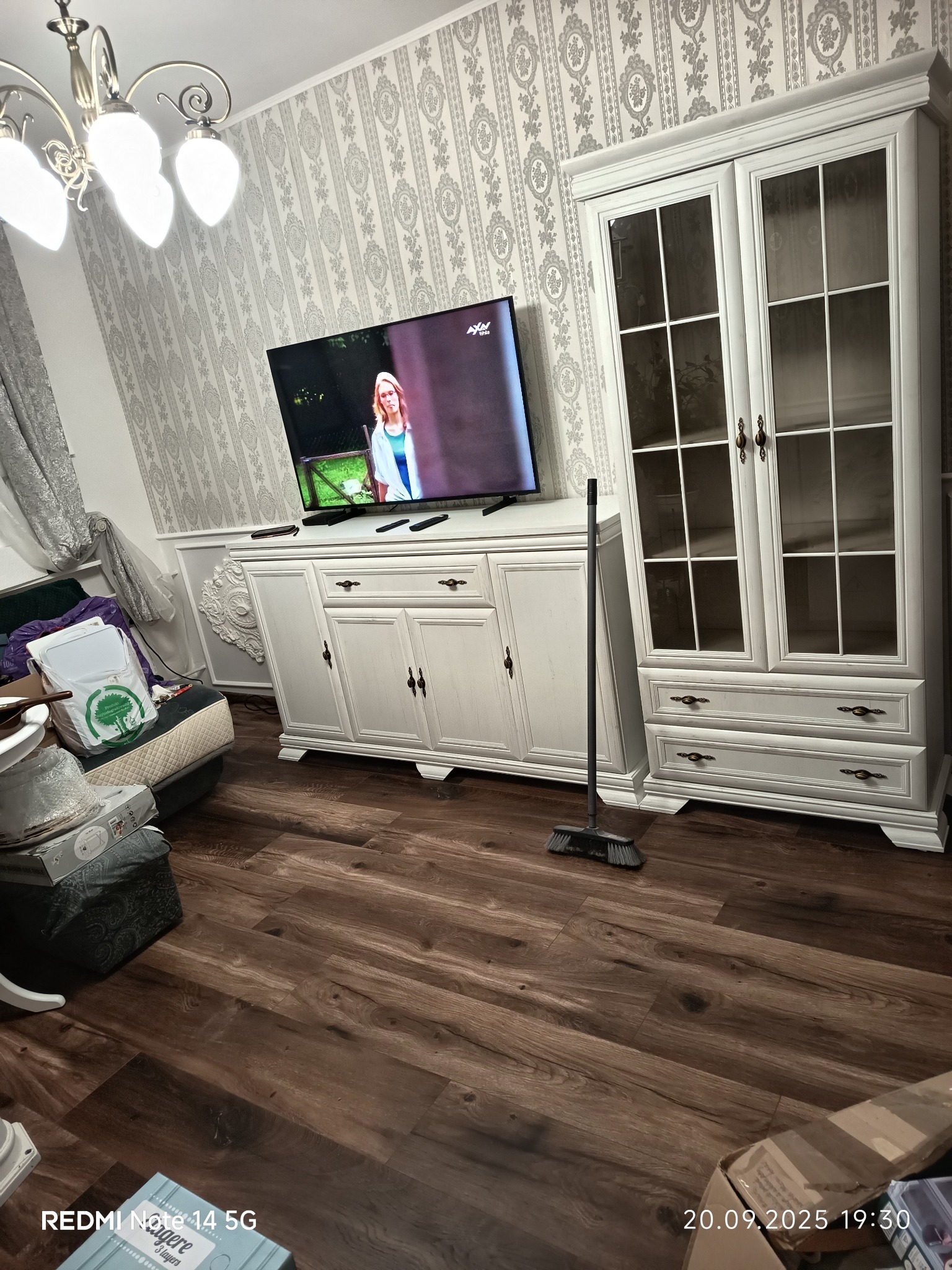 Jasne, odnowione wnętrze z drewnianą podłogą, białymi meblami i wzorzystą tapetą. Telewizor w tle. Świeżo po remoncie, ale jeszcze nie do końca posprzątane.