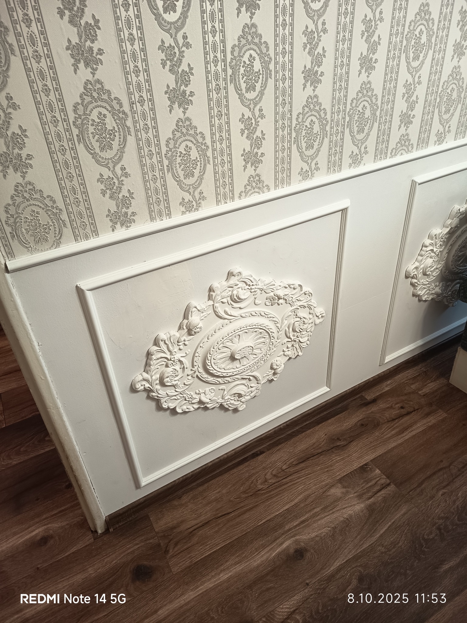 Eleganckie wykończenie ściany z białymi panelami i dekoracyjnymi sztukateriami, uzupełnione tapetą w subtelne, kwiatowe wzory oraz ciemnymi panelami podłogowymi.