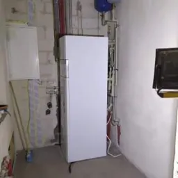 Biały piec gazowy kondensacyjny z dwoma zbiornikami wyrównawczymi na ścianie, w surowym pomieszczeniu z widocznymi instalacjami i elementami wykończeniowymi.