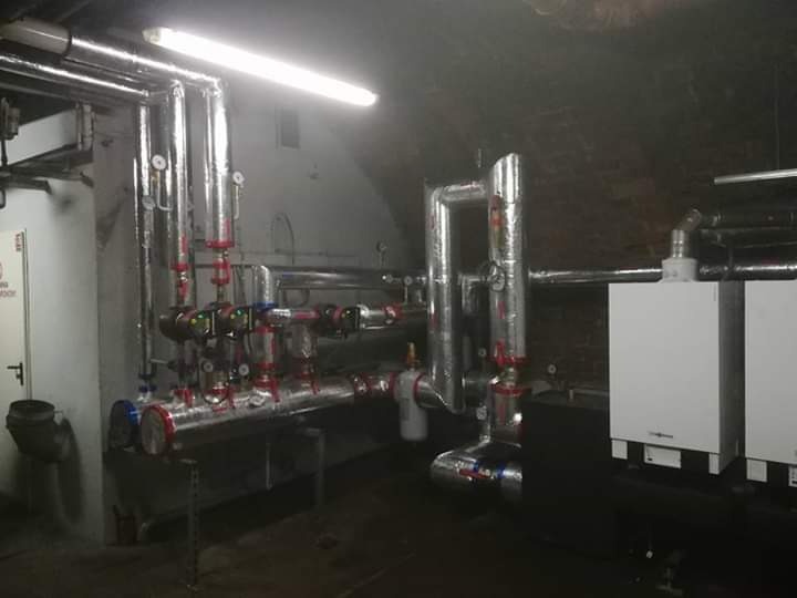 Złożony system rur z izolacją termiczną, zaworami i manometrami w kotłowni w Toruniu, z dwoma białymi piecami gazowymi Viessmann w tle.