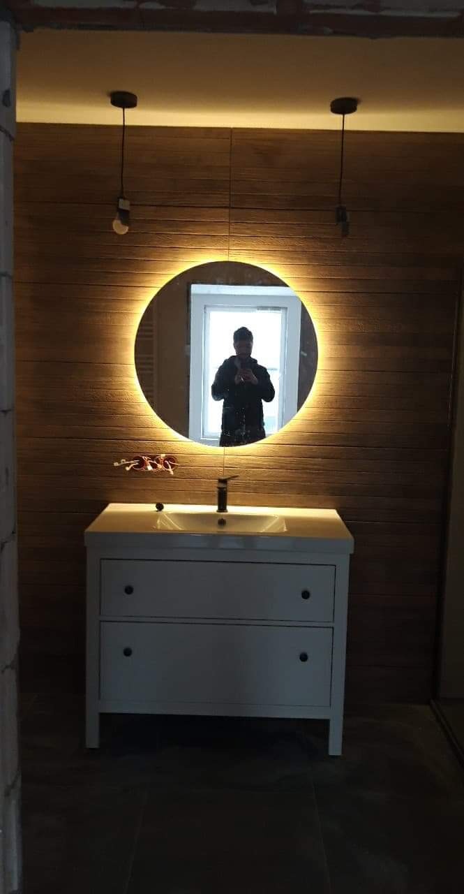 Nowoczesna łazienka z okrągłym lustrem podświetlanym LED, drewnopodobnymi panelami ściennymi i białą szafką pod umywalką, widoczne niedokończone wykończenia.