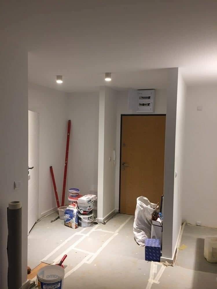 Korytarz w trakcie prac wykończeniowych: widoczne puszki z farbą, poziomica oparta o ścianę, podłoga zabezpieczona taśmą malarską, przygotowania do malowania ścian.