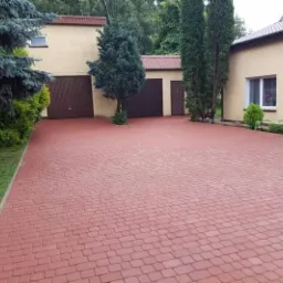 Czerwona kostka brukowa na podjeździe do garażu w domu jednorodzinnym, z widocznym trawnikiem i iglakami po bokach.