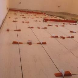 Usługi Ogólnobudowlane Jacek Kaczmarczyk - Układanie jasnych płytek podłogowych z użyciem systemu poziomowania, widoczne pomarańczowe kliny i czarne krzyżyki dystansowe.